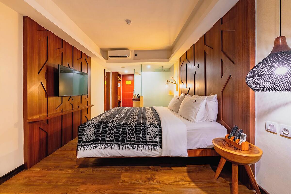 A hotel room with a large bed, a TV mounted on the wall, and a wooden nightstand with remote controls.