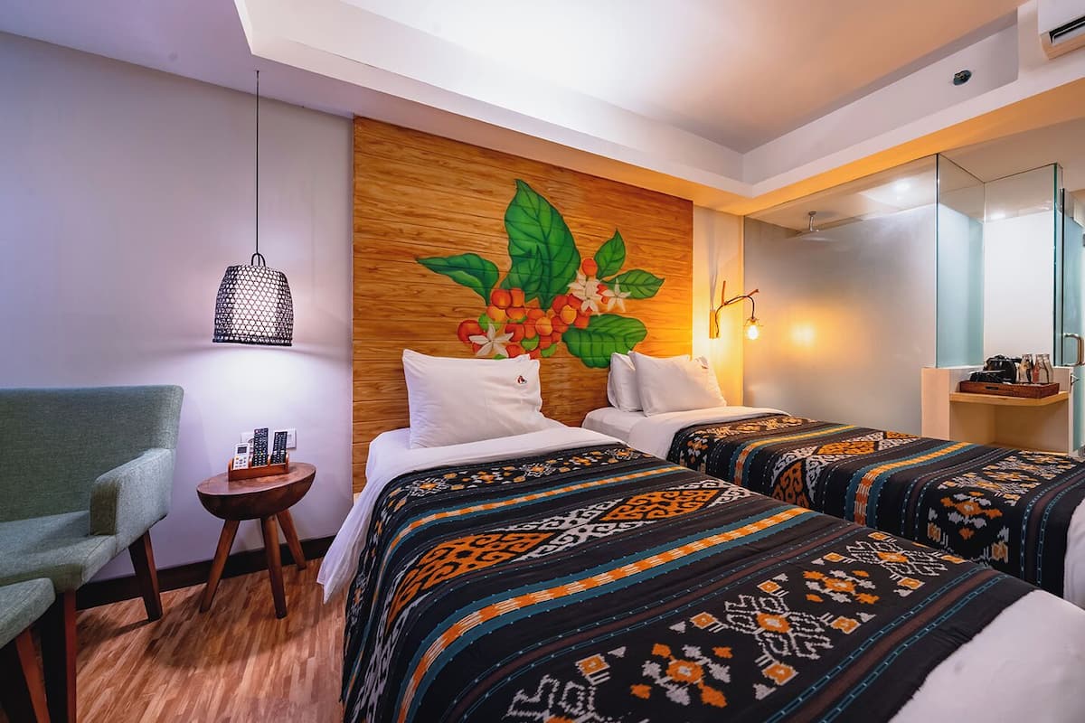 A hotel room with two beds, a green armchair, a small round table with a phone, and a wall with a floral mural.