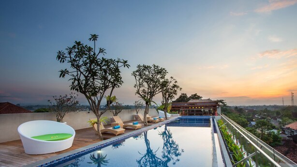 A rooftop infinity pool with lounge chairs and a white wicker chair, surrounded by trees and overlooking a cityscape at sunset. - MaxOne Hotels at Ubud (Ubud)