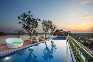 A rooftop infinity pool with lounge chairs and a white wicker chair, surrounded by trees and overlooking a cityscape at sunset.