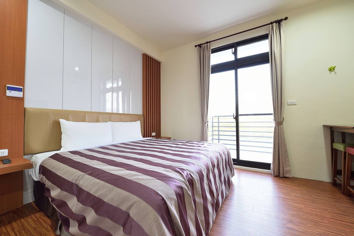 A hotel room with a large bed, wooden flooring, a desk, and a window with curtains.