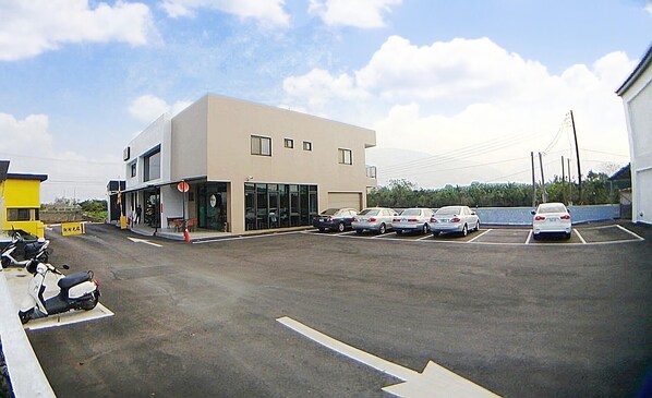 A parking lot with several cars and a motorcycle, a building with large windows and a flat roof, and a clear sky. - CoCo Motel (Puli)