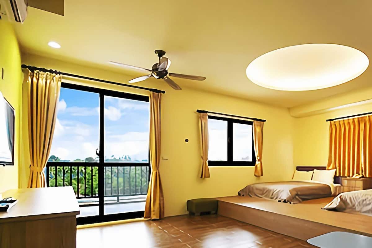 A hotel room with a balcony, a ceiling fan, a bed, a desk, and a view of greenery.