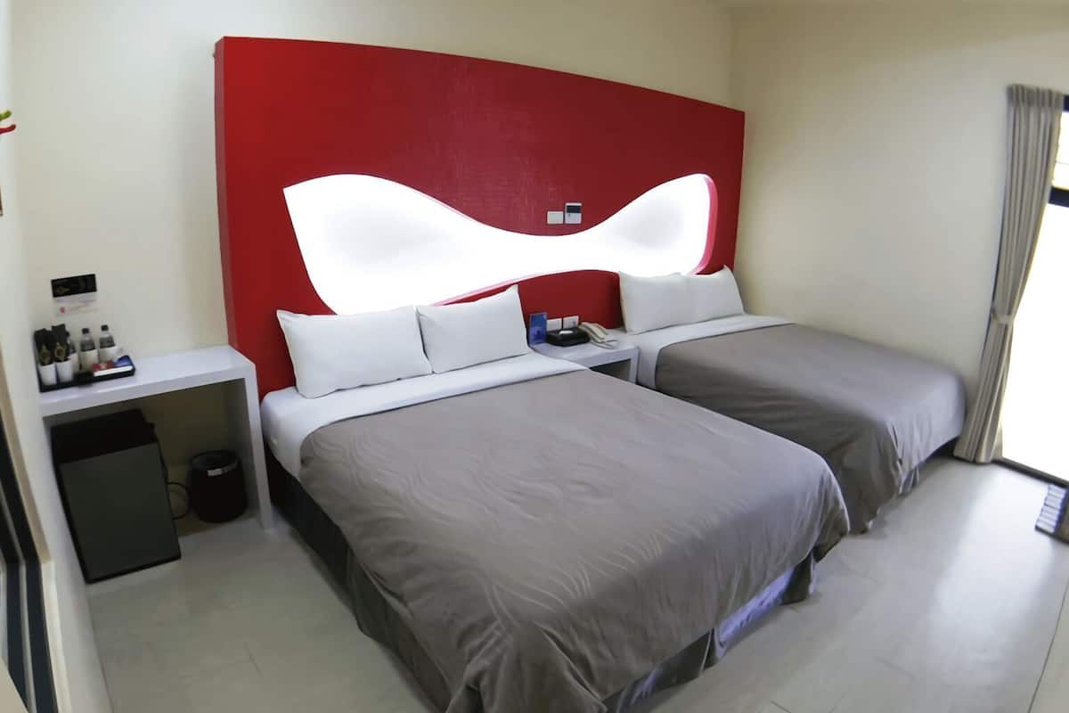 A hotel room with two beds, a red accent wall, a bedside table with a phone, and a small trash bin.