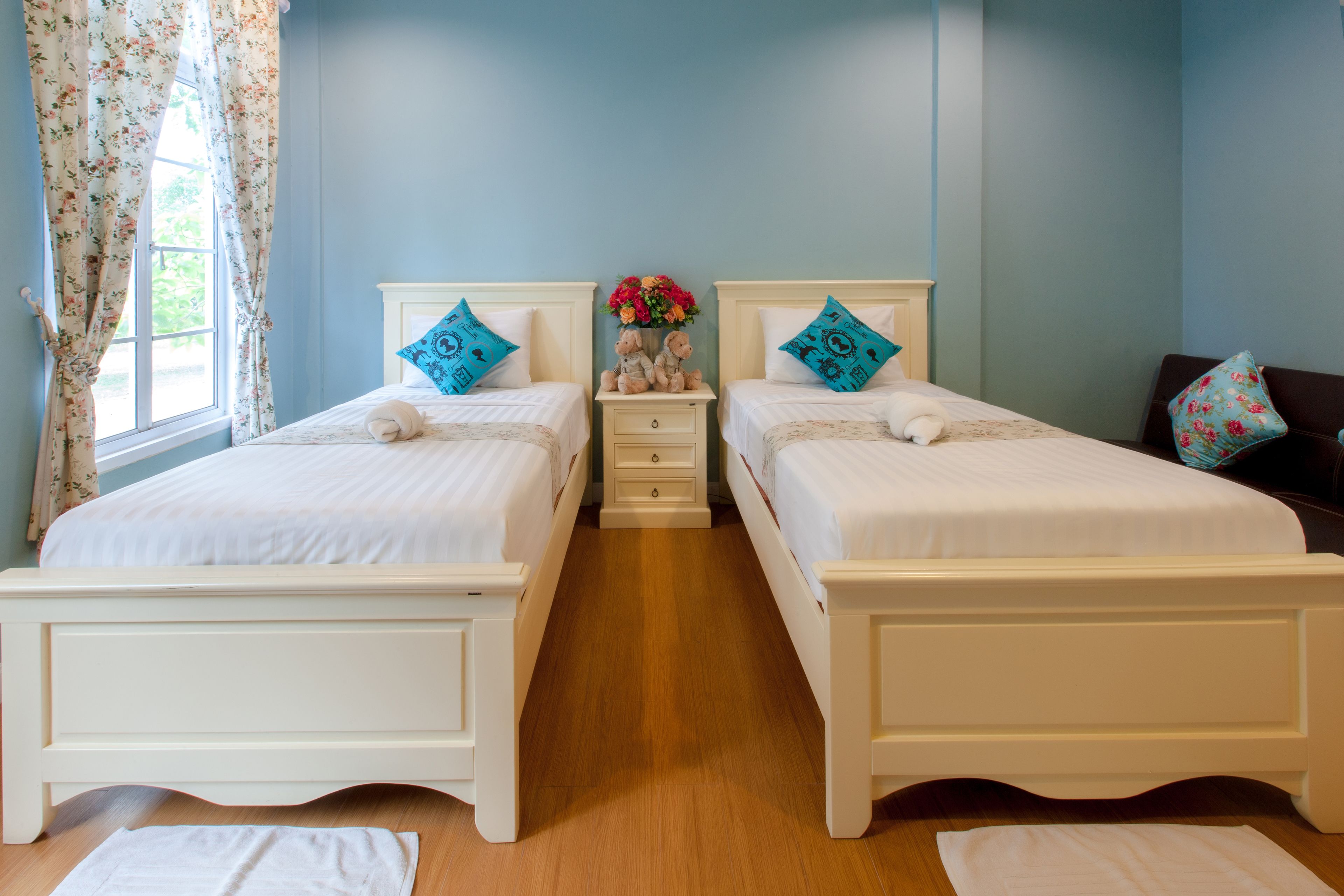 A double room with two beds, a nightstand with a vase of flowers, and a window with floral curtains.