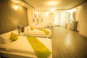 A hotel room with two beds, a desk, and a chair. There is a small table with a vase of flowers and a picture frame. The room has a wooden floor and a white wall with a light fixture. - New Hotel & Apartment (Ho Chi Minh City)