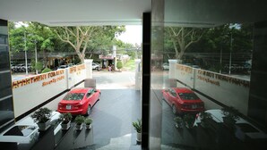 Vista desde un pasillo de hotel, con dos coches rojos estacionados afuera, un cartel de servicio de seguridad las 24 horas y el reflejo de los alrededores en la pared de cristal.