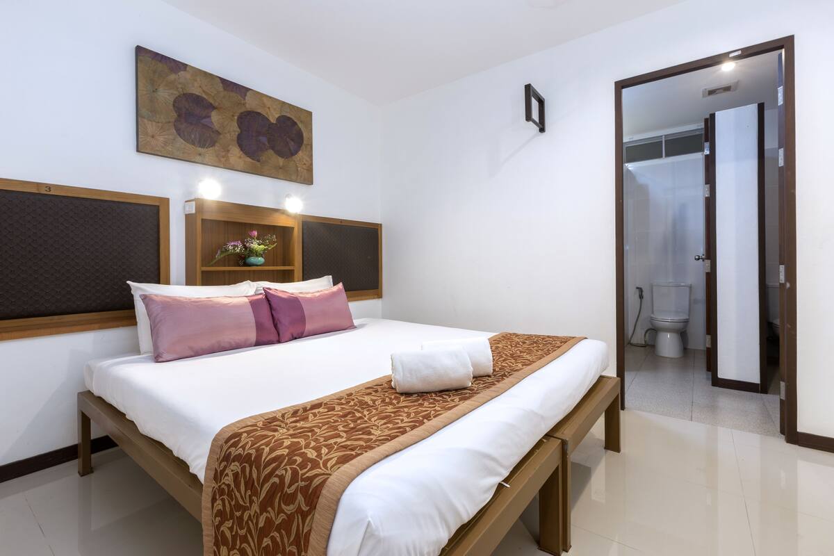 A hotel room with a bed, a wooden headboard, a bedside table, a mirror, and a bathroom visible through an open door.