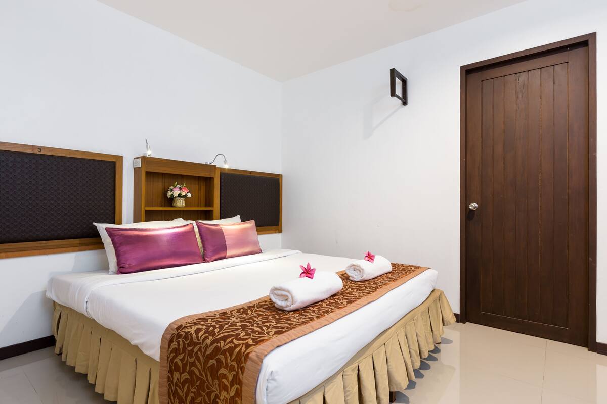 A hotel room with a bed, a wooden door, and a small wooden shelf with a vase.