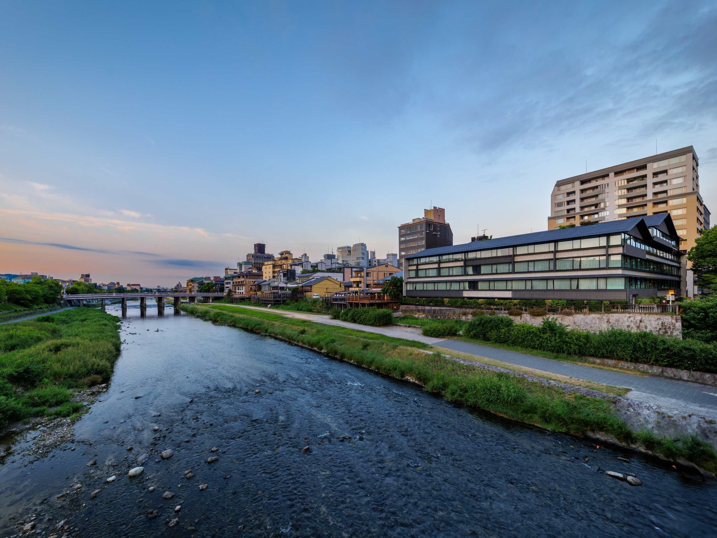 A riverside view of a cityscape with modern buildings, a bridge, and a pathway alongside the river.