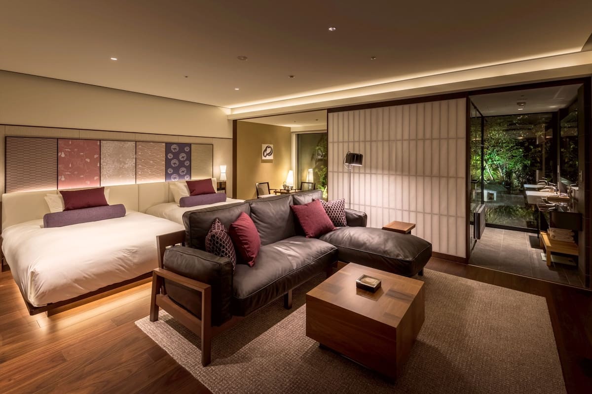 A modern hotel room with a large bed, a sofa, a coffee table, and a view of the garden through a sliding door.