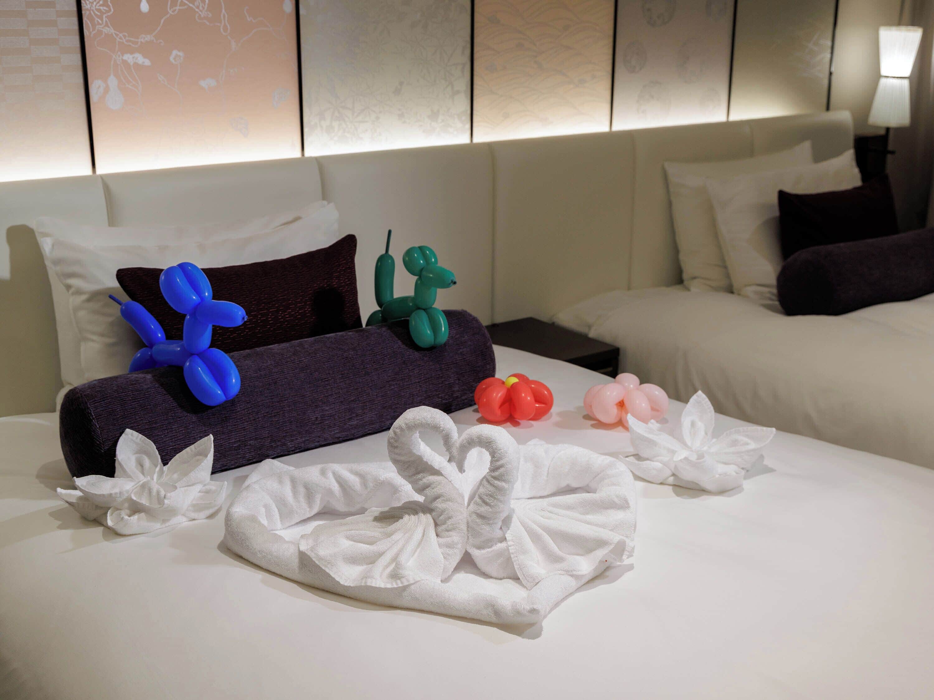 A hotel room with a neatly made bed, pillows, and decorative items including a blue and green balloon sculpture and a red and pink balloon flower.