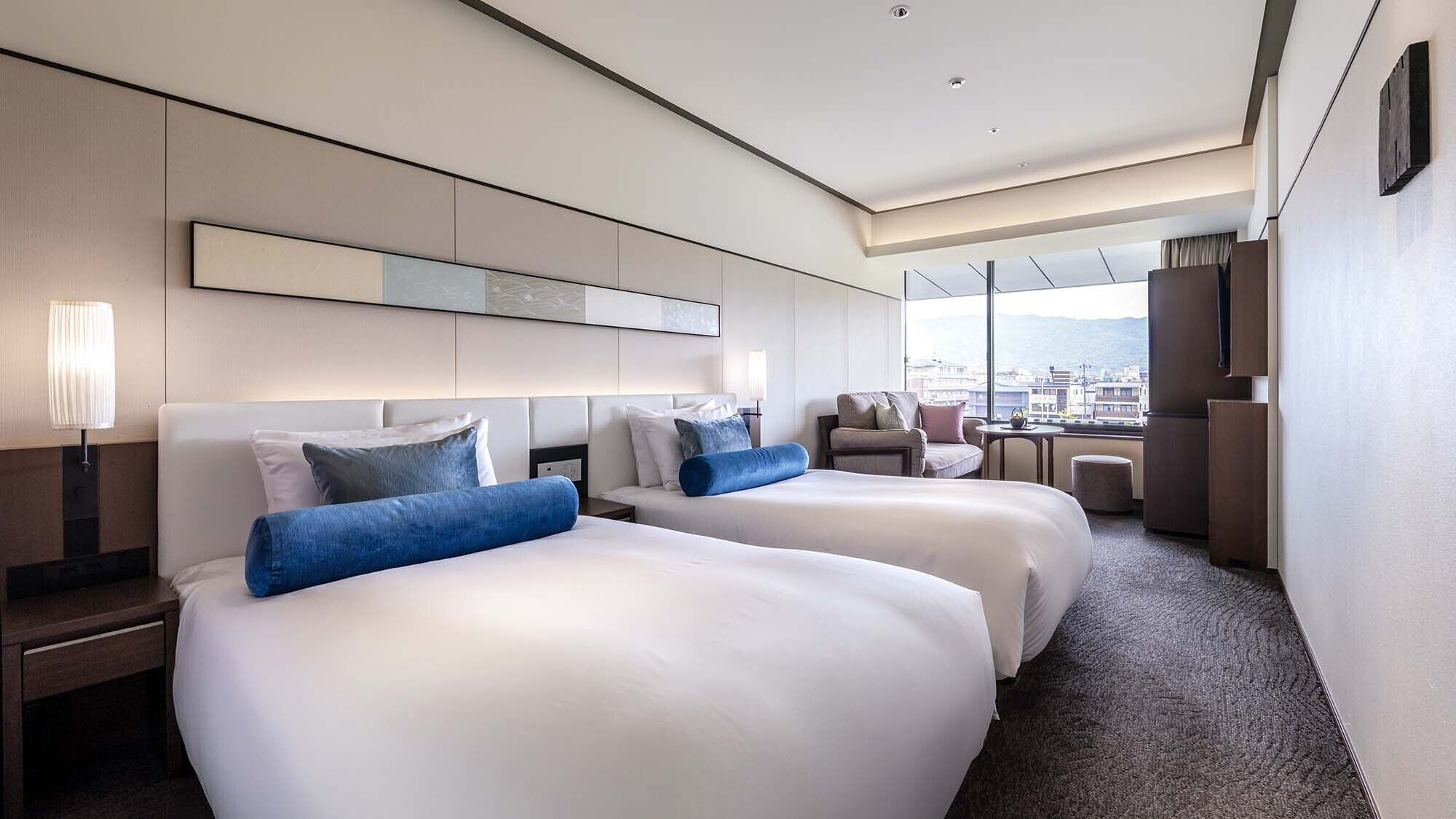 Ein Hotelzimmer mit zwei Betten, einem Sofa, einem Schreibtisch und einem großen Fenster mit Blick auf die Stadt.