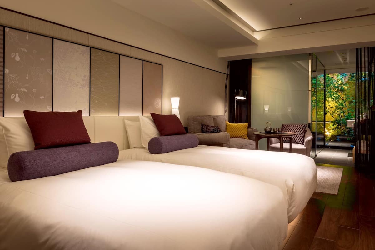 A modern hotel room with two large beds, a sofa, a small table, and a view of greenery through a glass door.