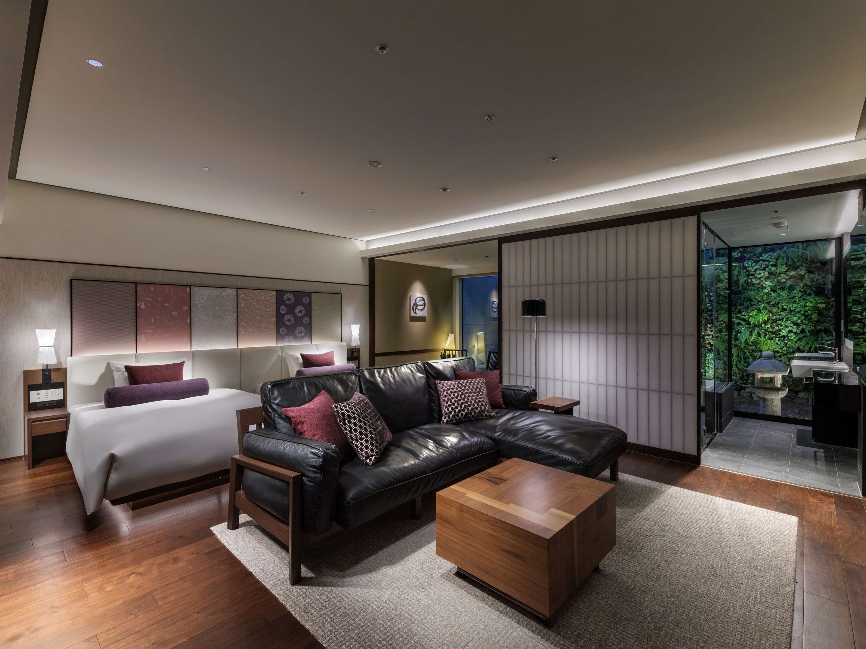 A modern hotel room with a large bed, a leather sofa, a wooden coffee table, and a sliding glass door leading to a bathroom with a stone sink.