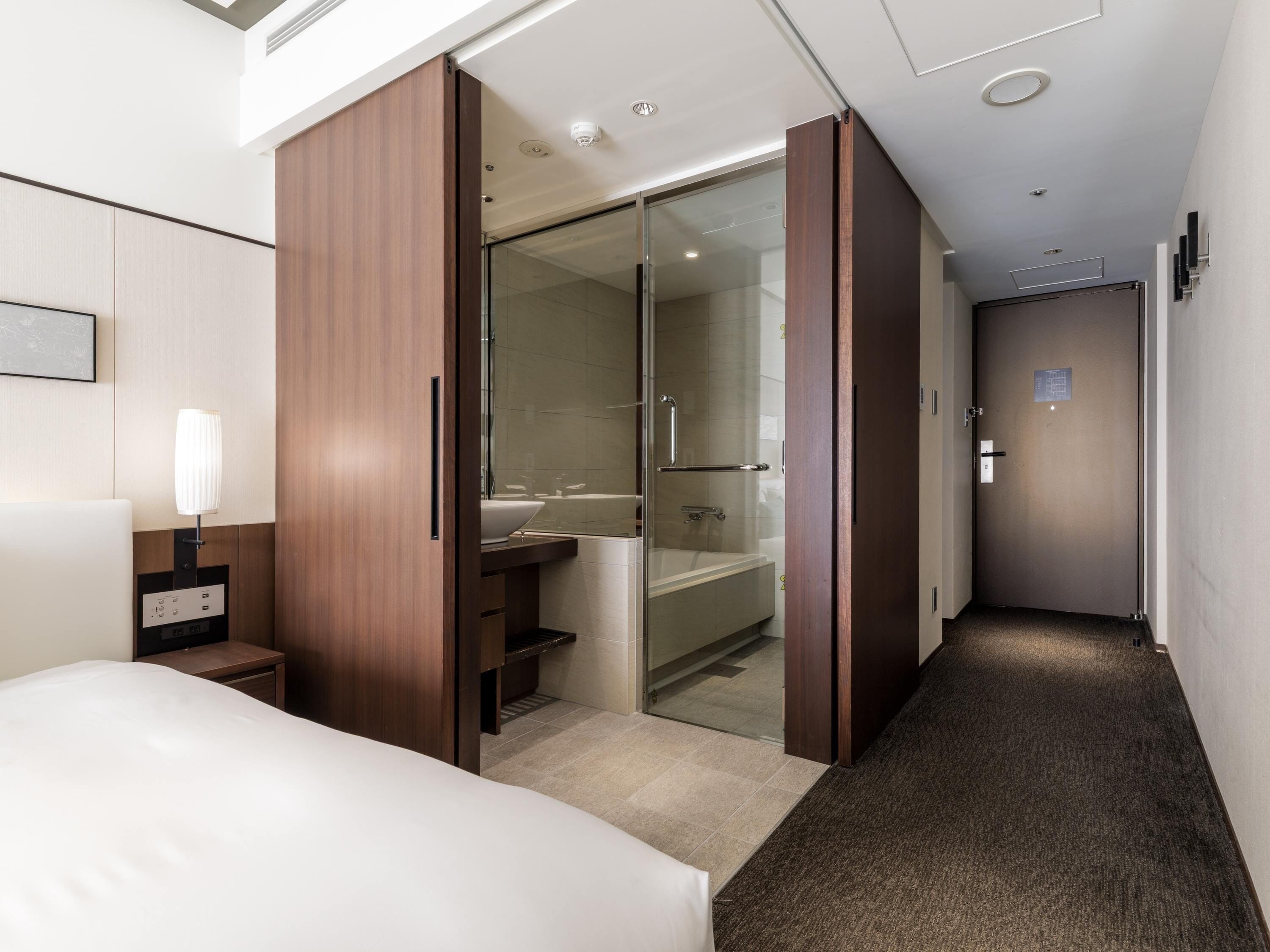 A hotel room with a bed, a desk with a lamp, a wardrobe, a bathroom with a shower, and a door leading to another room.