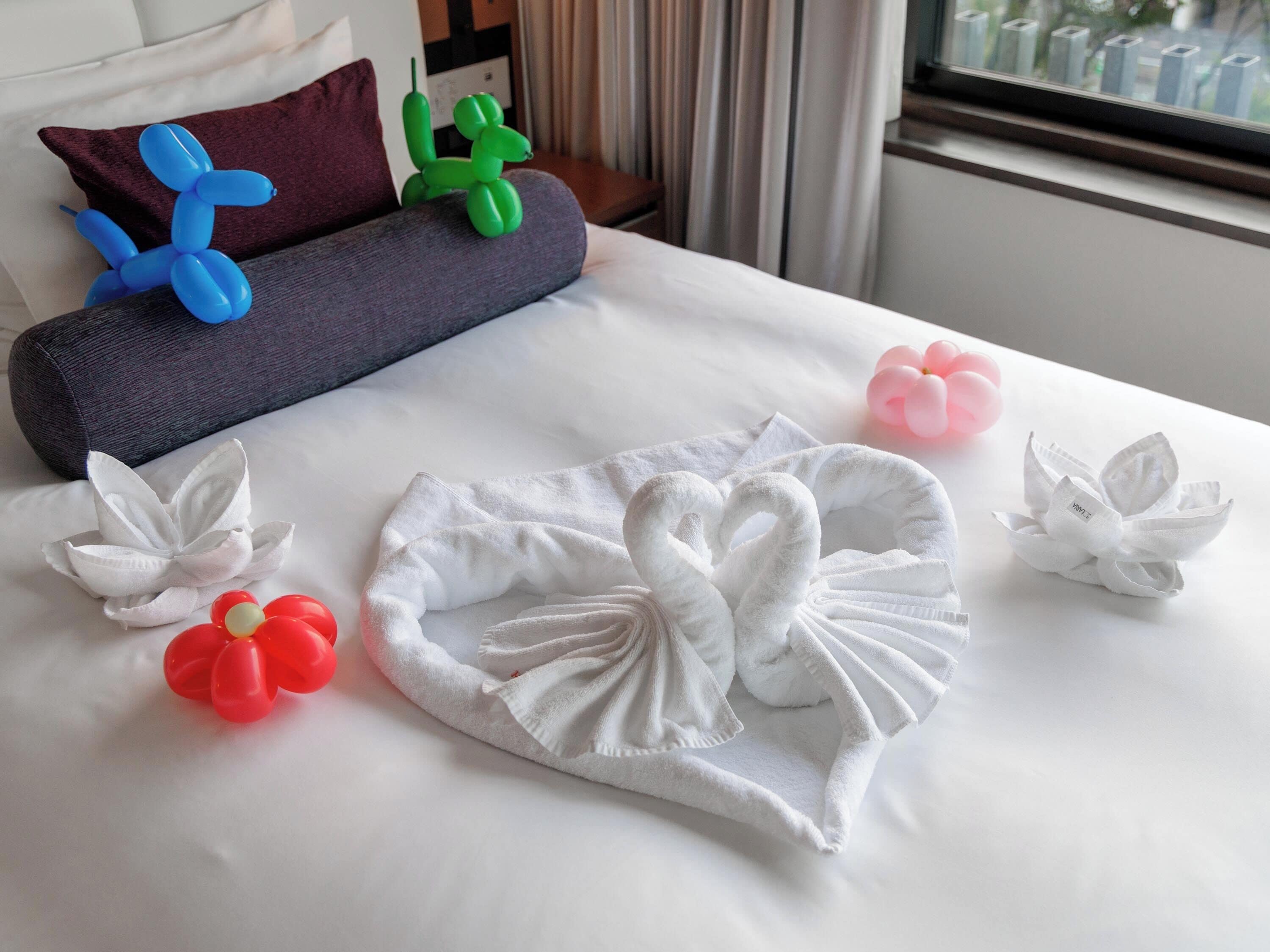 A neatly made bed with white towels arranged in swan and flower shapes, a gray pillow, and colorful balloon sculptures.