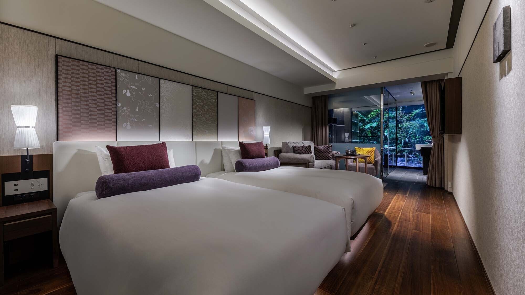 Ein Hotelzimmer mit zwei Betten, einem Schreibtisch, einem Stuhl und Blick auf einen Wald durch eine Glasdoor.