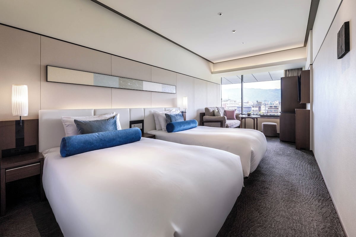 A hotel room with two beds, a desk, a chair, and a view of the city.