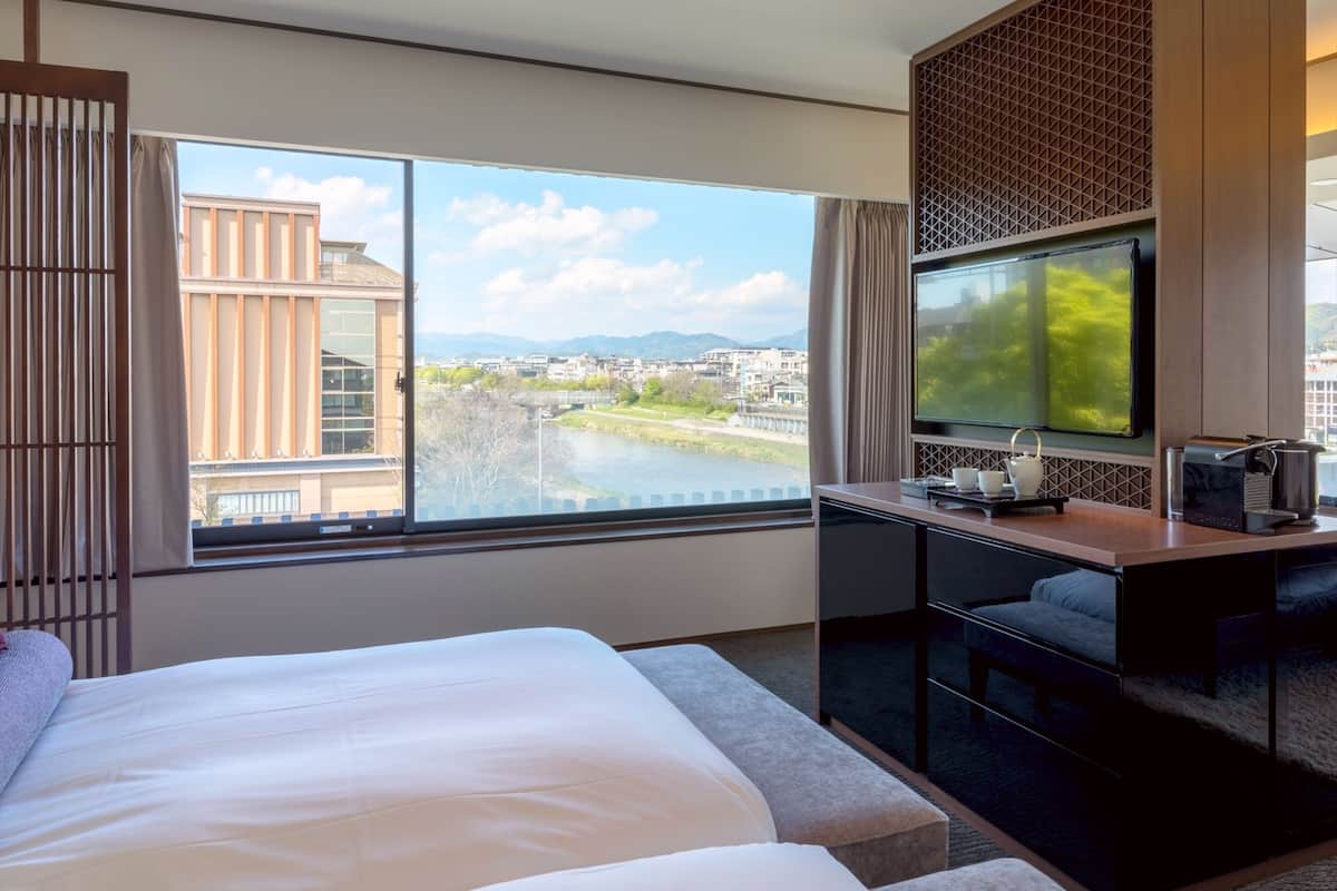 A hotel room with a large bed, a desk with a TV, and a view of a river and buildings.