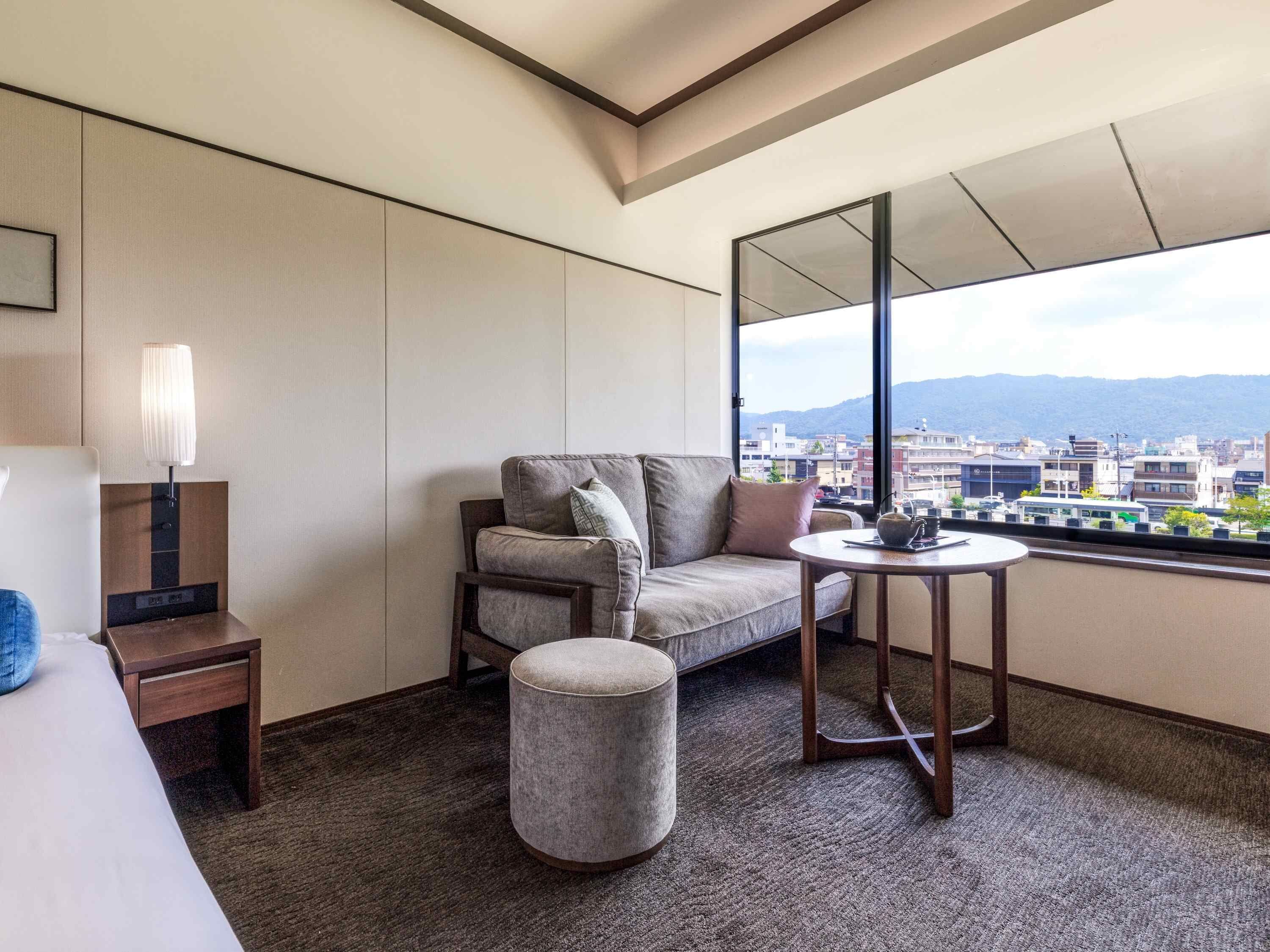 A hotel room with a sofa, a small table, a lamp, and a view of the city through large windows.