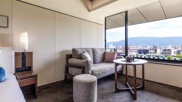 A hotel room with a sofa, a small table, a lamp, and a view of the city through large windows.