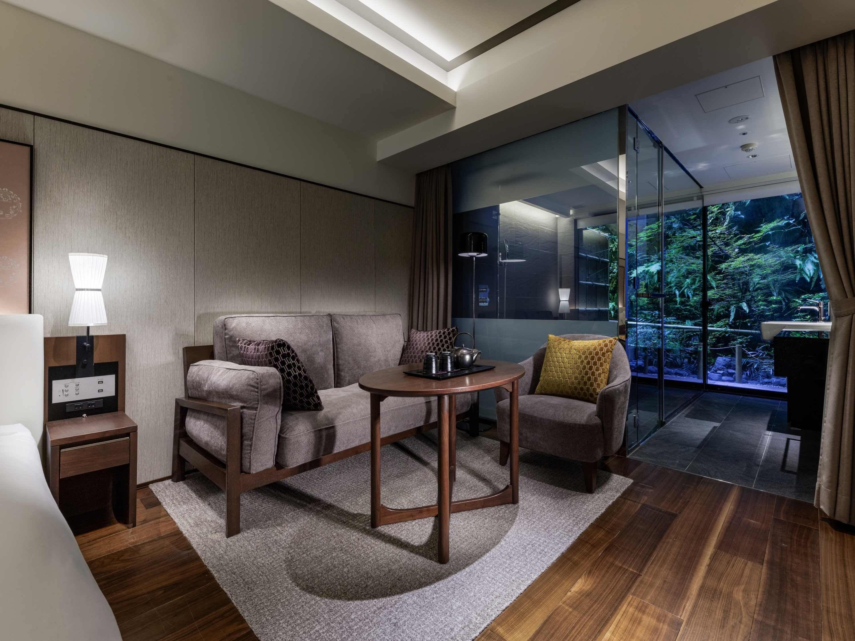 A modern hotel room with a sofa, armchair, and a small table. There is a nightstand with a lamp, a large window with a view of greenery, and a bathroom visible through the glass wall.