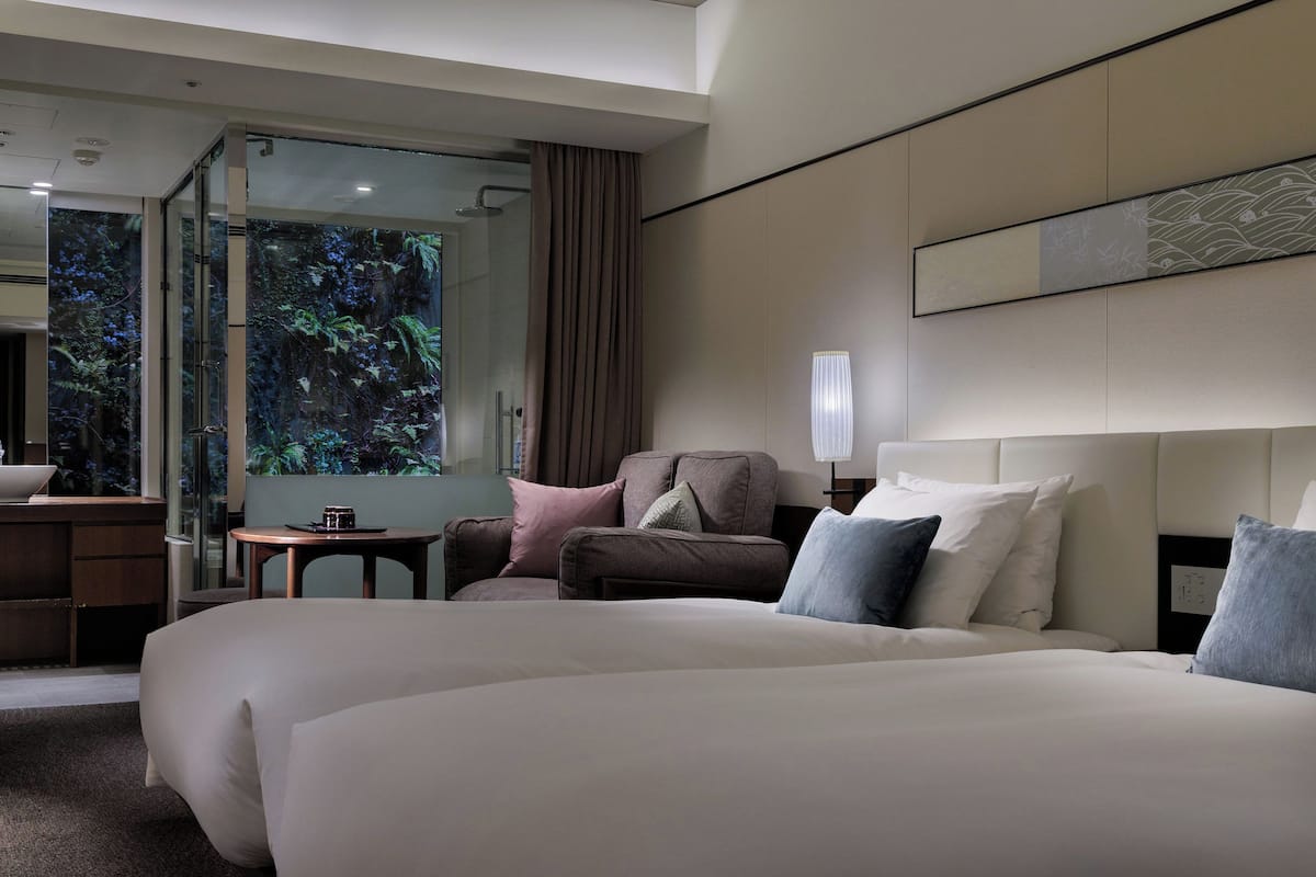 A hotel room with two large beds, a sitting area with a chair and a small table, and a large window with a view of greenery.