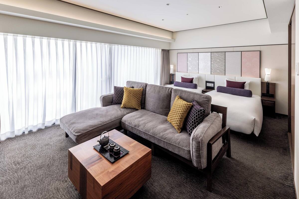 A modern hotel room with a sectional sofa, a single bed, a wooden coffee table, and a large window with sheer curtains.