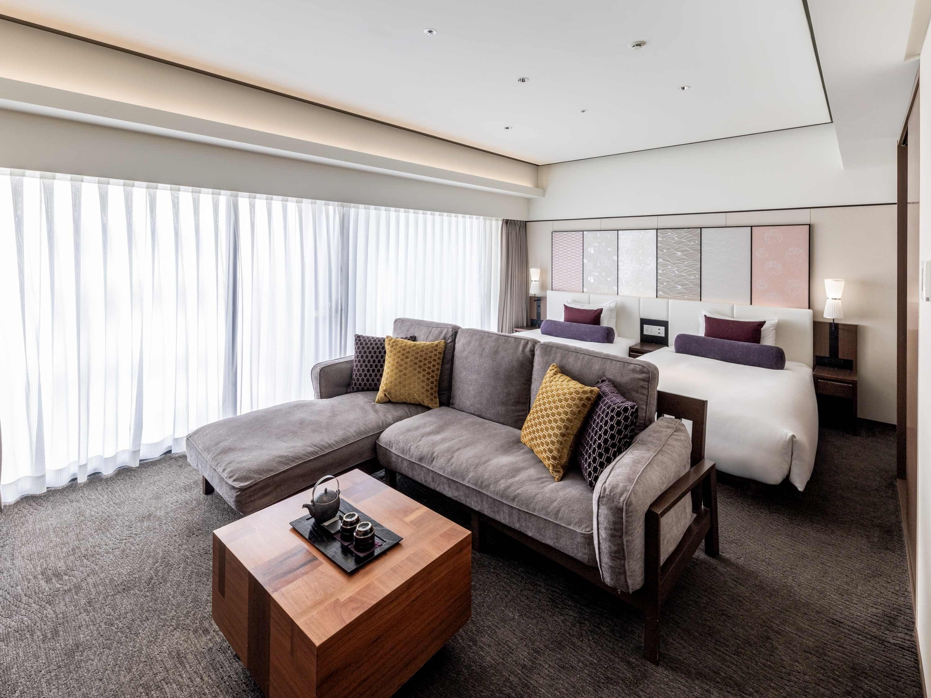 Una habitación de hotel moderna con un sofá modular, una cama individual, una mesa de centro de madera y un ventanal con cortinas translúcidas.
