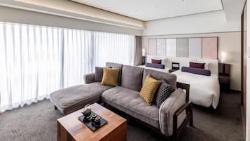 Una habitación de hotel moderna con un sofá modular, una cama individual, una mesa de centro de madera y un ventanal con cortinas translúcidas.