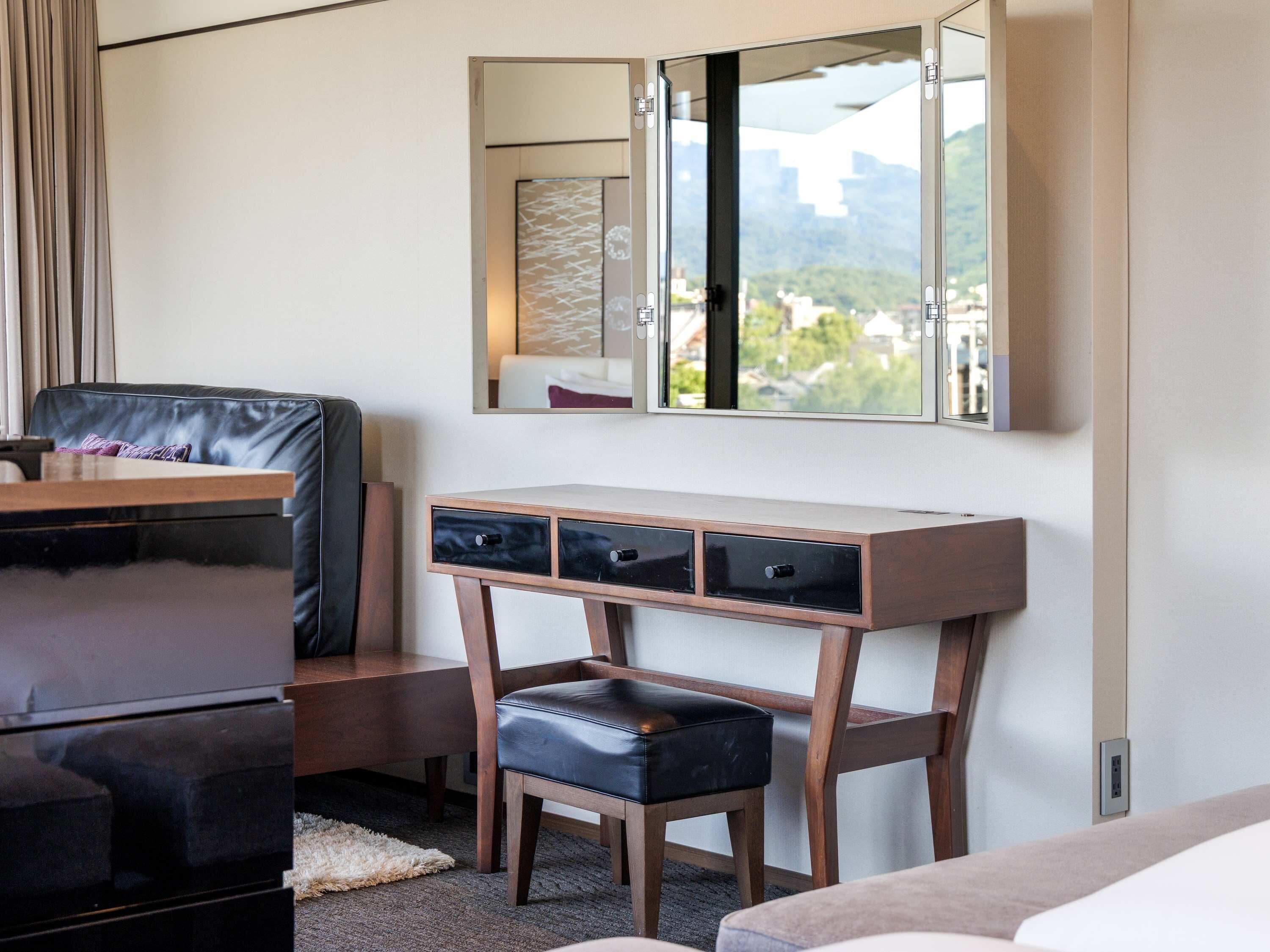 A modern hotel room with a wooden desk, a black leather chair, a small stool, and a large mirror above a window.