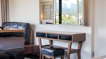 A modern hotel room with a wooden desk, a black leather chair, a small stool, and a large mirror above a window.