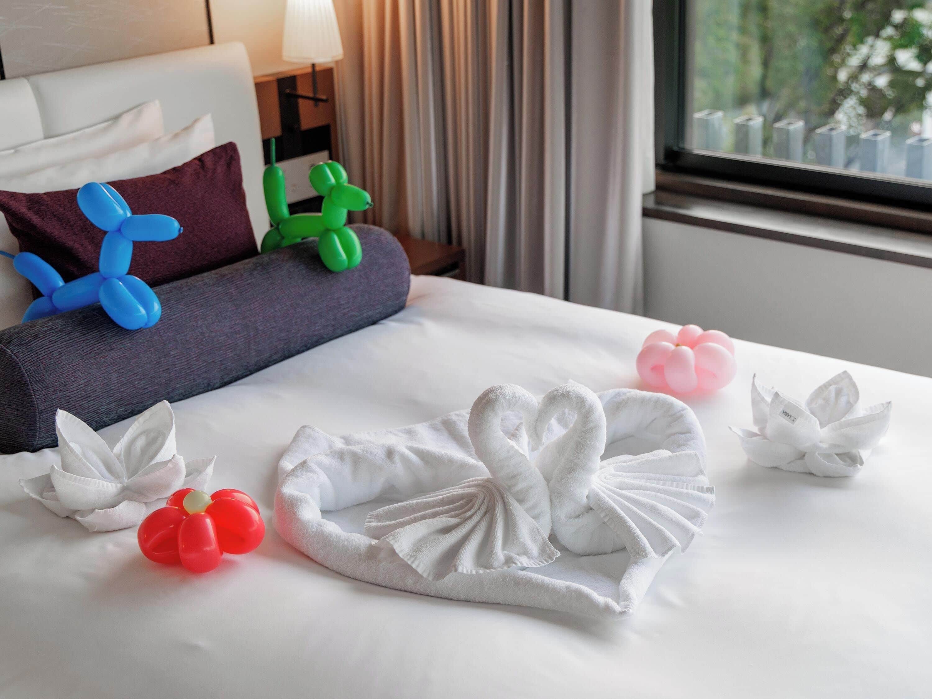 Ein ordentlich bezogenes Bett mit einem weißem Handtuch in Schwanenform, roten und grünen Ballons sowie einer weißen Blumenkomposition.