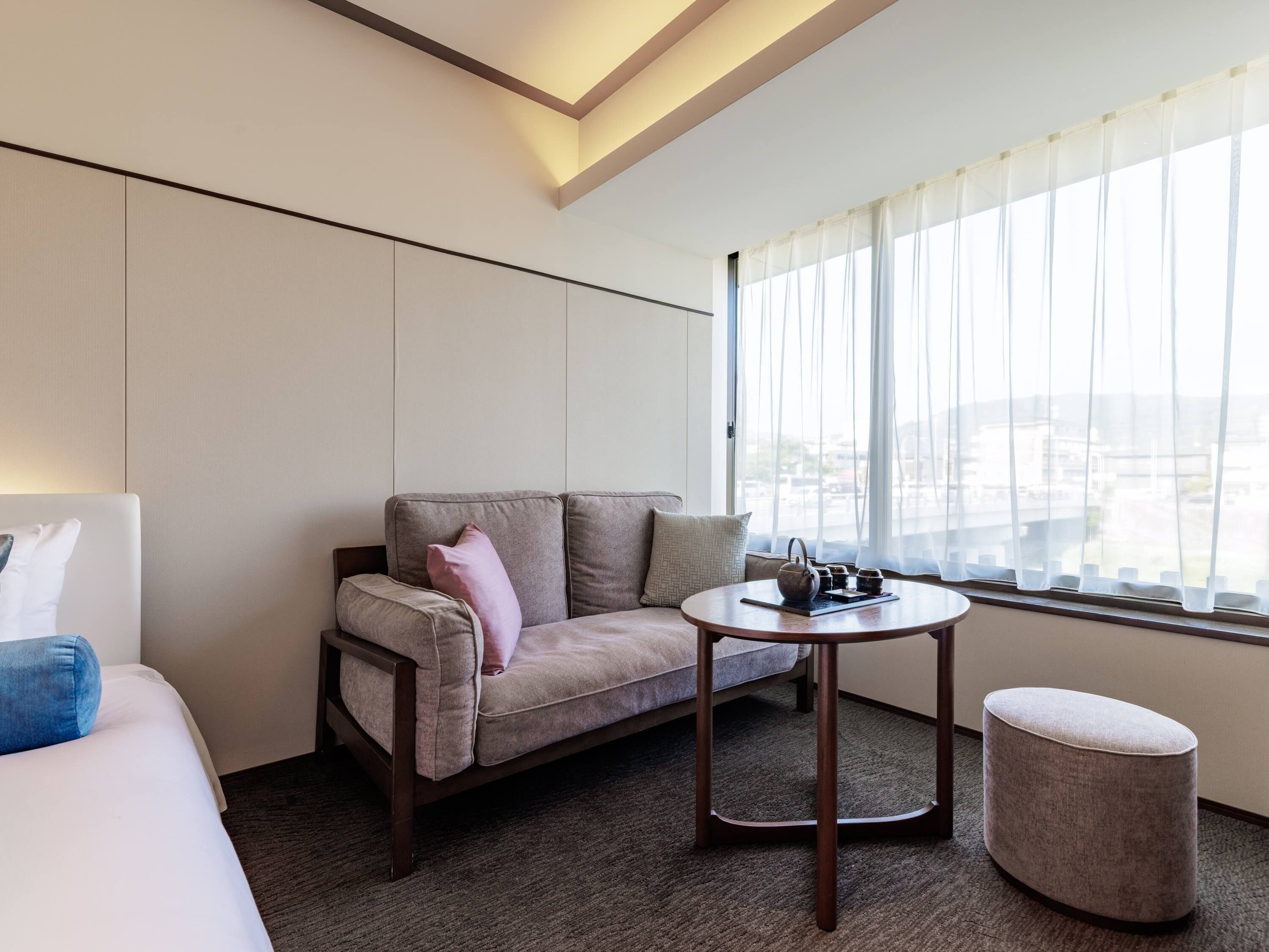 A modern hotel room with a sofa, a small round table with a teapot, and a view of the city through sheer curtains.