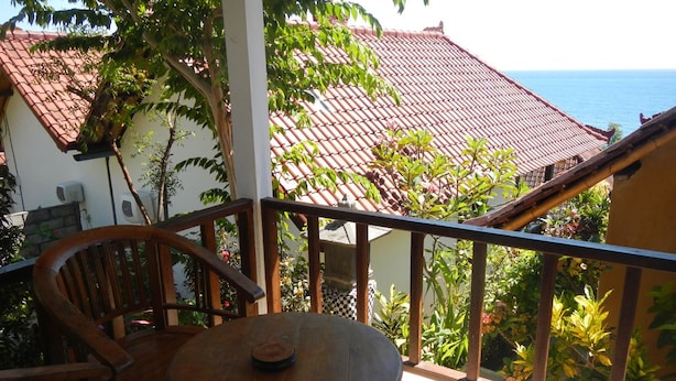 Varanda de madeira com mesa e cadeiras, com vista para um telhado de telhas vermelhas e o mar.