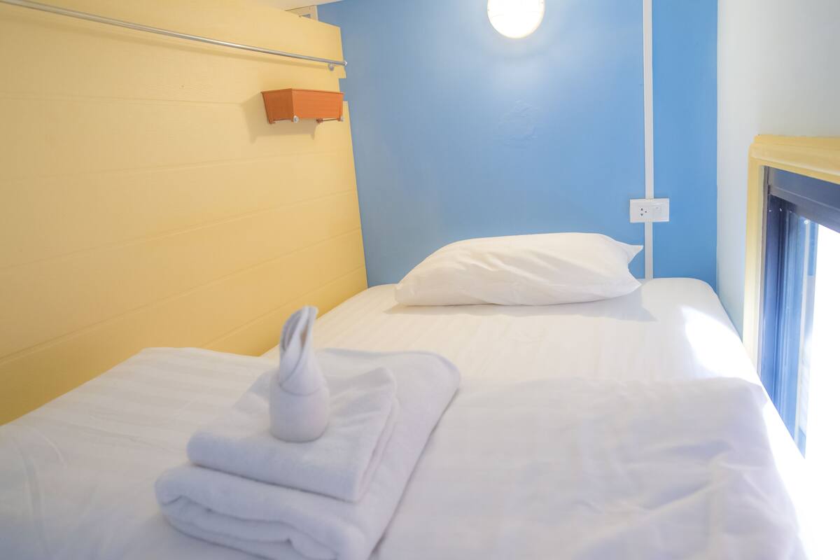 Une chambre d’hôtel avec un lit, du linge de lit blanc, une serviette pliée, une table de chevet et une fenêtre avec des rideaux.