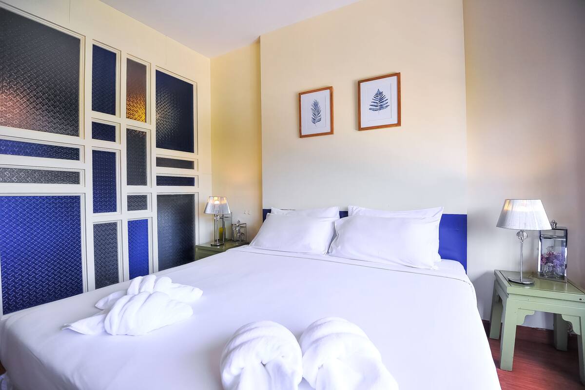 Une chambre d’hôtel avec un grand lit, du linge de lit blanc, deux œuvres d’art représentant des feuilles encadrées au mur, et une table de chevet avec une lampe et un récipient en verre.