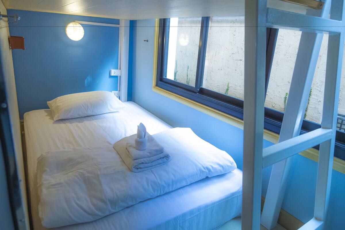 Un lit avec du linge de lit blanc et une serviette pliée, une fenêtre donnant sur l’extérieur et un mur bleu.
