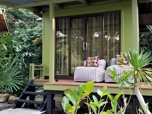 A covered outdoor seating area with a wooden deck, cushioned chairs, and a large window with curtains.