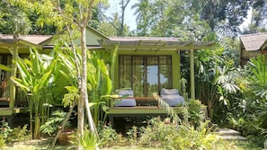 A green-roofed building with a covered porch, two cushioned seating areas, and lush tropical vegetation.