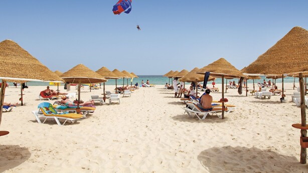 A beach with sun loungers and umbrellas, a clear sky, and a person parasailing.