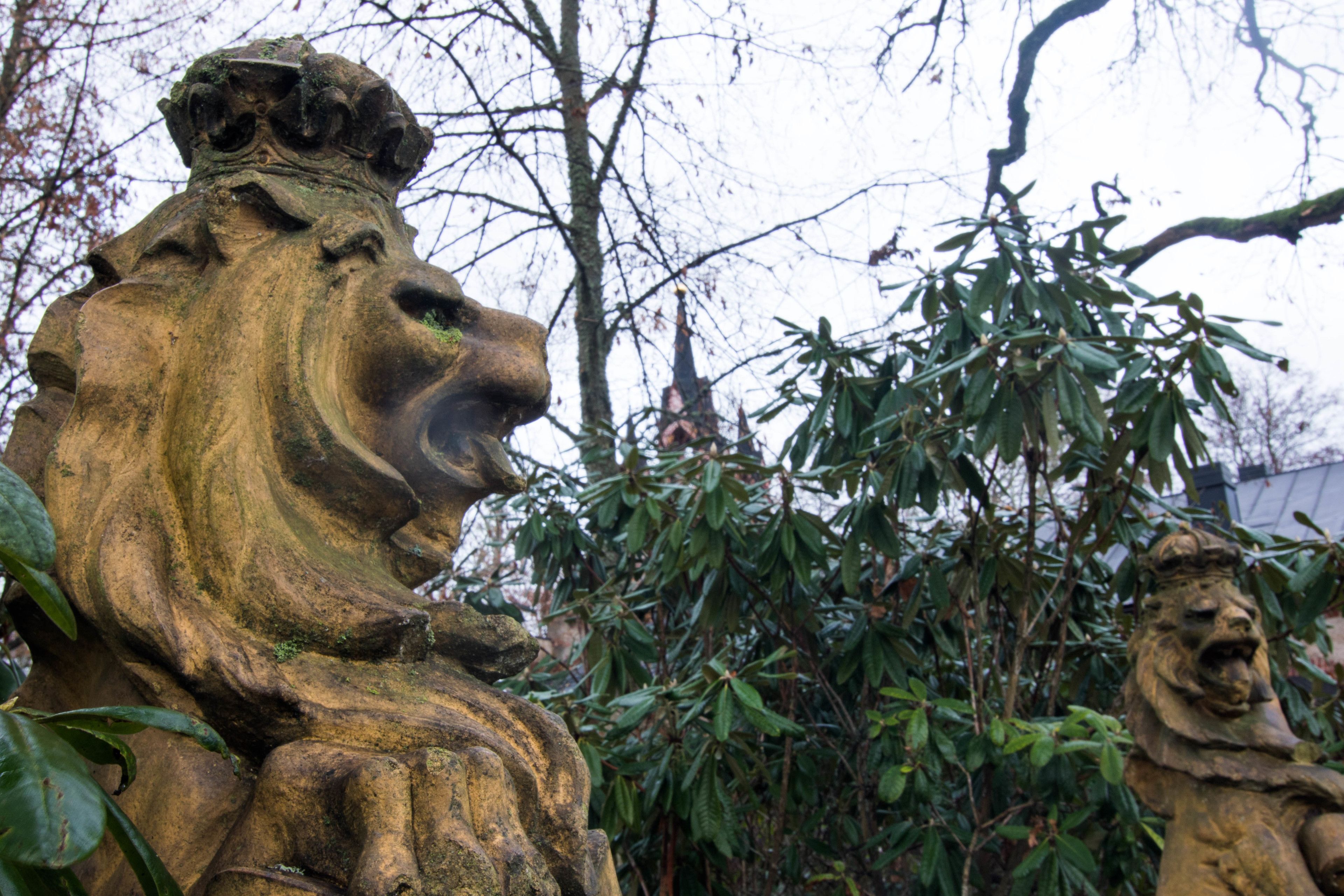 A stone lion statue in a garden with trees and a building in the background.