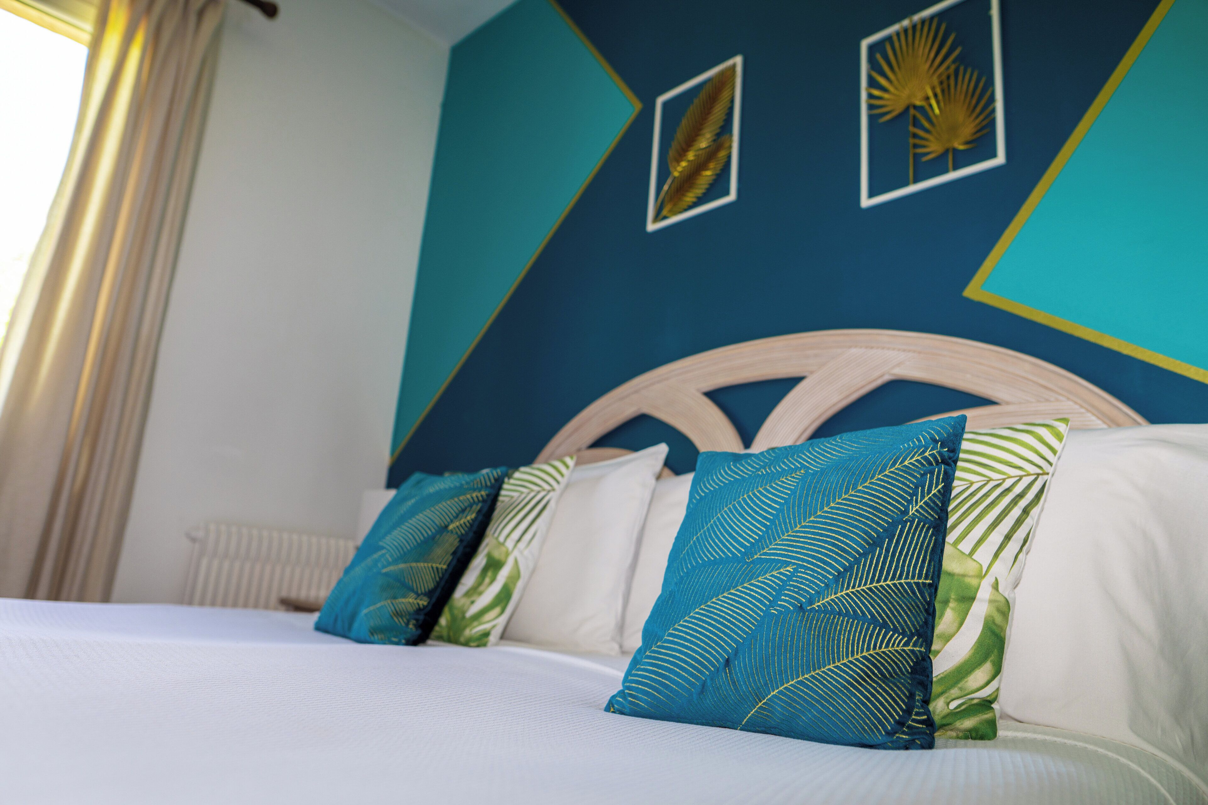 Un lit avec une literie blanche et des coussins décoratifs, dans une pièce aux murs de couleur turquoise et ornée d’œuvres d’art murales aux motifs géométriques.