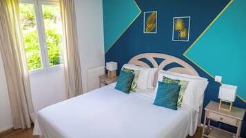 Una cama bien hecha con ropa blanca y almohadas estampadas en azul, una mesita de noche con lámpara, y una ventana con cortinas.