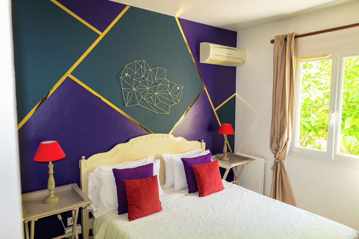 Un dormitorio con una cama, dos mesitas de noche, un aire acondicionado instalado en la pared y una ventana con cortinas.