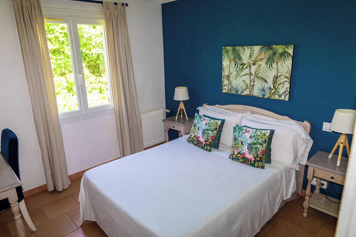 Un dormitorio con cama, dos mesitas de noche, una silla, ventana con cortinas, radiador y un cuadro en la pared.