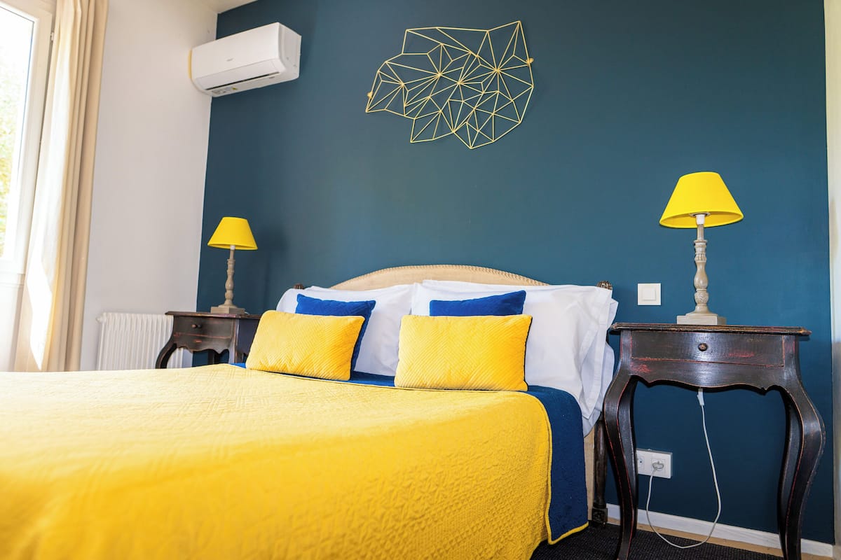 Una cama bien hecha con colcha amarilla y azul, dos lámparas de noche, un adorno geométrico en la pared y un equipo de aire acondicionado.