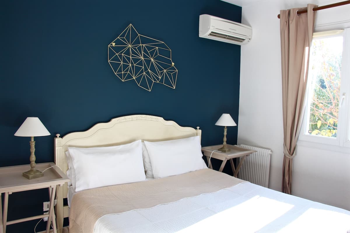 Un dormitorio con una cama, dos mesitas de noche con lámparas, un adorno geométrico en la pared, un aire acondicionado y una ventana con cortinas.