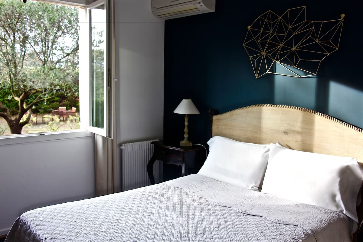 Un dormitorio con cama, lámpara, decoración geométrica en la pared y ventana con vista a árboles.
